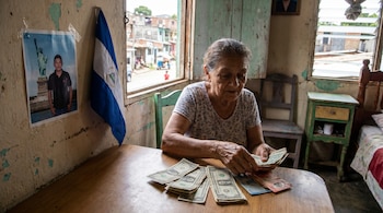 Anciana sentada a una mesa de madera contando dólares y córdobas. En la pared, foto de joven con la Estatua de la Libertad y bandera de Nicaragua. Ventana al fondo.