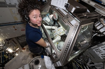 La astronauta Jessica Meir, con cabello rizado y auriculares, sonríe mientras manipula jeringas dentro de una caja de guantes en la Estación Espacial Internacional