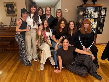 Diez mujeres sonríen a cámara en una sala con piso de madera, un piano, un cuadro y un mueble antiguo con adornos religiosos