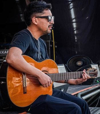 Un hombre con gafas de sol y camiseta negra toca una guitarra de madera en un escenario