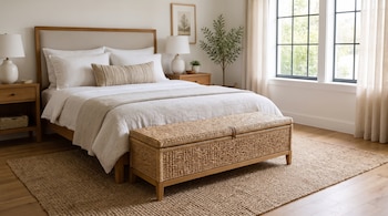 Una cama matrimonial con cojines, un baúl de mimbre a los pies, dos lámparas sobre mesas de madera y una ventana con cortinas.