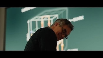 Hombre de cabello gris con gafas y jersey oscuro, sonríe. Fondo verde azulado borroso con un diagrama de cubo y estructuras anaranjadas