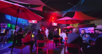 Personas sentadas a la mesa y de pie en una terraza de noche bajo sombrillas e iluminación de color rojo y azul
