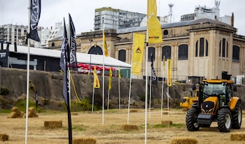 Campo con un tractor amarillo, fardos de heno, banderas publicitarias, un gran edificio de piedra y edificios urbanos al fondo