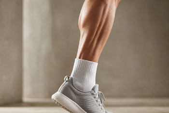 Plano cercano de una pierna con calcetín blanco y zapatilla gris, apoyada de puntillas con los músculos de la pantorrilla en tensión.