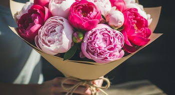 Primer plano de un ramo de peonías rosas y fucsias, envuelto en papel kraft con un lazo de cuerda, sostenido por manos en un entorno de luz natural.