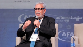 Hombre de cabello y barba gris, con gafas, traje oscuro y corbata azul, hablando en un micrófono y gesticulando. Sentado frente a una pantalla LED azul
