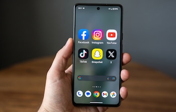 Mano humana sostiene un teléfono móvil con pantalla mostrando iconos de Facebook, Instagram, YouTube, TikTok, Snapchat y X, junto a barra de búsqueda de Google