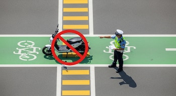 Agente de tránsito con uniforme azul y amarillo apunta a una motocicleta eléctrica en ciclovía, con un símbolo de prohibición encima, sobre asfalto gris.