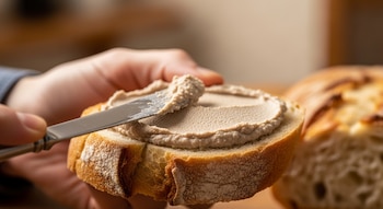Primer plano de una mano sosteniendo una rodaja de pan dorado y untando paté de hígado cremoso con un cuchillo de manteca.