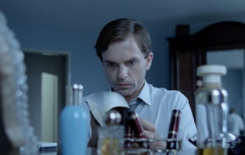 Un hombre con camisa blanca y corbata observa objetos. En primer plano, botellas de vidrio, un soporte metálico y objetos rojos