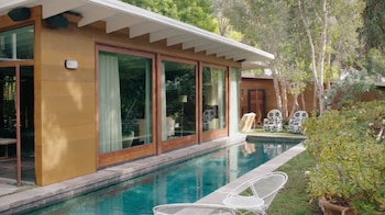 Vista exterior de una casa moderna de color madera con grandes puertas de vidrio, una piscina larga y estrecha, césped y árboles verdes bajo el sol