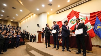 Homens uniformizados recebem prêmios e medalhas na bandeira peruana e no campo da bandeira azul. Muitas pessoas na plateia aplaudem no auditório.