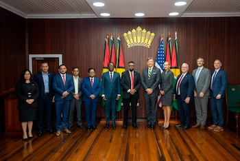 Doce personas, once hombres y una mujer, posan de pie en una sala con paredes de madera, banderas de Guyana, bandera de Estados Unidos y un escudo dorado