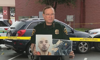 Hombre en uniforme de sheriff con gafas, sosteniendo dos fotos: una de un hombre y otra de un arma dentro de un vehículo. Fondo con autos y cinta policial