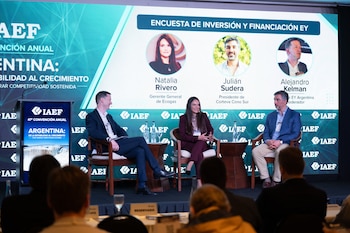 Tres personas sentadas en un escenario conversan durante una presentación frente a una pantalla gigante con sus fotografías y nombres