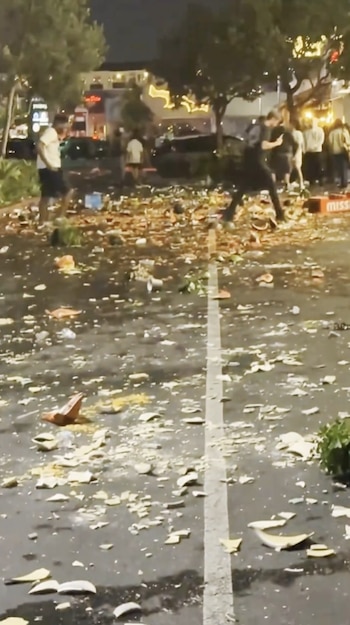 Calle con una línea divisoria blanca, cubierta de basura y escombros. Varias personas de pie al fondo en una zona iluminada