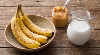 Tres bananas maduras en un plato hondo, un frasco de vidrio con pasta de maní y una jarra con leche de coco sobre una mesa de madera.
