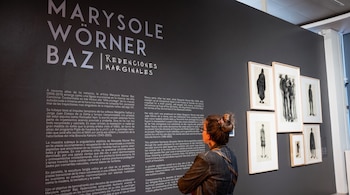 Pared oscura con texto blanco de exposición "Marysole Wörner Baz", persona de espaldas mirando cuadros enmarcados con dibujos, iluminación de galería.