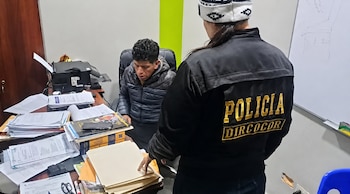 Un hombre sentado frente a un escritorio lleno de documentos es inspeccionado por una agente con la inscripción Policía DIRCOCOR en la espalda