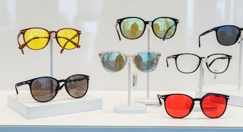 Siete pares de gafas exhibidas en un escaparate blanco, con lentes espejadas, transparentes, amarillas y rojas, y monturas de distintos colores.