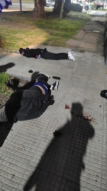 Vista aérea de dos personas vestidas de oscuro, boca abajo sobre una acera gris cuadriculada, con una mancha de sangre roja cerca