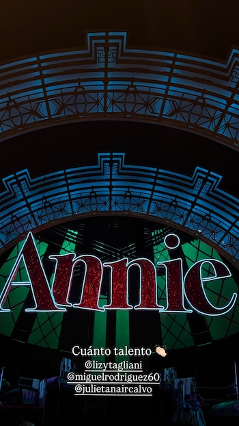 Vista frontal del cartel luminoso rojo con el texto "Annie" y un borde de neón azul, sobre un escenario oscuro con elementos azules y camas