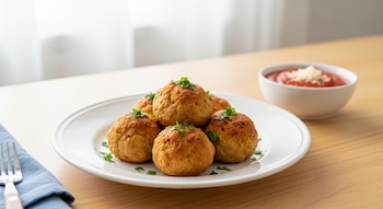 Plato blanco con seis albóndigas de pollo doradas decoradas con perejil, al lado un bol con salsa de tomate y queso, sobre una mesa de madera.