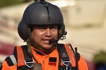 Un hombre con casco gris, micrófono en la barbilla, traje naranja y chaleco salvavidas negro y naranja con el logo CMC