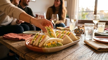 Una mano toma un sándwich de una bandeja sobre una mesa de madera con vasos y copas en una reunión.