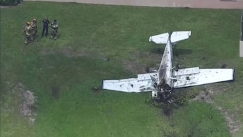 Vista aérea de una avioneta blanca estrellada y parcialmente quemada en un campo de hierba verde, con bomberos y personal de emergencia cerca