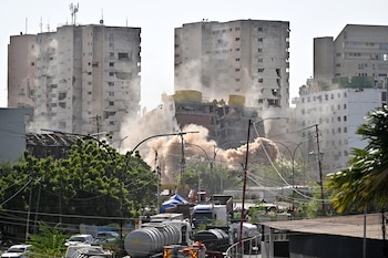 Una demolición genera una nube de polvo densa entre edificios altos. Hay postes de luz, cables eléctricos, árboles y vehículos en primer plano