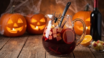 Jarra de vidrio con bebida roja y fruta sobre mesa de madera, junto a una botella de vino y tres calabazas talladas con velas encendidas.