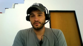Hombre barbudo con gorra y auriculares con micrófono, mirando al frente. Se observa una escalera blanca detrás y un marco naranja lateral