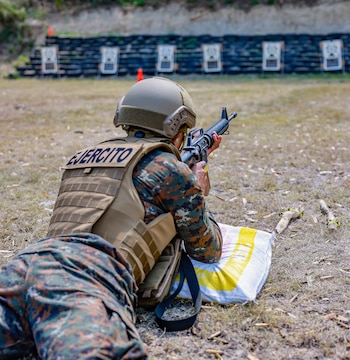 Soldado con uniforme camuflado, chaleco y casco, en posición prona apuntando un rifle hacia blancos en un campo de tiro. Vainas de bala en el suelo