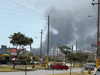 Una avenida urbana con automóviles, árboles y postes de electricidad bajo una gran columna de humo oscuro en el fondo