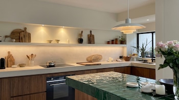 Vista de una cocina moderna con gabinetes de madera oscura, encimeras claras, y una isla con superficie de azulejos verdes, adornada con utensilios y plantas.