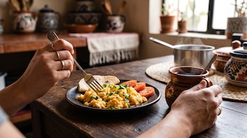 Manos sobre una mesa de madera sostienen un tenedor y un jarro de barro junto a un plato con huevos revueltos, tomate y una tortilla.