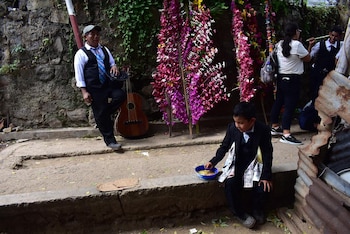 Hombre con guitarra y niño sentado con dinero en su traje, junto a estructuras florales moradas y blancas, en un entorno de pared de piedra y tierra