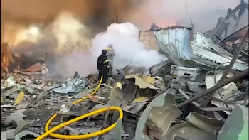 Dos bomberos con equipo protector sofocan las llamas y el humo entre los restos de un edificio bombardeado, rodeados de escombros y metal retorcido