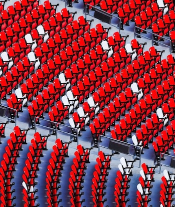 Vista aérea de numerosas filas de butacas de estadio en tonos rojo brillante y blanco, organizadas en un patrón diagonal sobre una superficie gris
