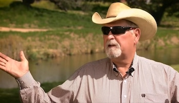 Un hombre de mediana edad con sombrero vaquero, gafas de sol, camisa a cuadros y barba canosa gesticula con su mano derecha frente a un estanque y vegetación