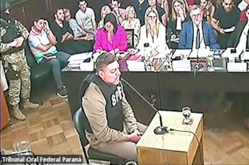 Hombre vestido con chaleco "SPF" sentado frente a un micrófono en un tribunal, con un guardia militar y otras personas presentes en la sala