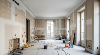 Interior de un piso vacío en Madrid en plena reforma, con paredes a medio alisar, techos altos, ventanas clásicas y materiales de construcción en el suelo.