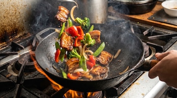 Un wok negro sobre una llama, salteando trozos de cerdo y verduras coloridas como brócoli, pimientos rojos y cebolla, con vapor elevándose.