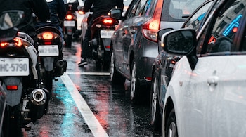Fila apretada de motocicletas y automóviles con luces de freno encendidas sobre asfalto mojado, se ven placas borrosas y líneas de calle.