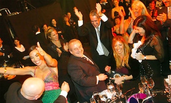 Grupo de personas sonriendo y bailando. Hombre con traje a rayas riendo, mujer con top colorido y falda verde bailando. Mesas con copas y botellas