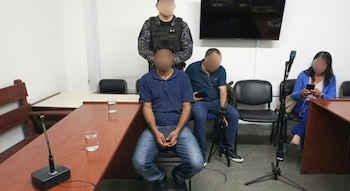 Un hombre sentado frente a un agente policial en una sala con sillas, mesas de madera, dos vasos de agua y un micrófono de pie