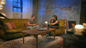 Dos hombres sentados en un sofá y un sillón, mesa de madera, botellas, vasos, discos de vinilo, lámparas, pared de ladrillo, cortina, ventana, alfombra