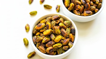 Dos tazones blancos llenos de pistachos sin cáscara y salados, con varios pistachos esparcidos sobre una superficie blanca lisa.